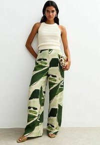 Wijdlopende broek in groen en beige met een bladenpatroon, gecombineerd met een witte gebreide haltertop en versierde sandalen, gehouden met een clutch tas.
