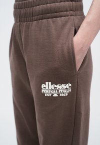 Brązowe dresy z prążkowanym pasem w talii i boczną kieszenią, z białym logo, które brzmi "ellesse Perugia Italia EST 1959" na udzie.