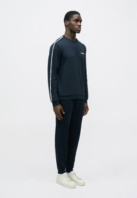 Muž stojaci v tmavomodrej teplákovej súprave s bielymi bočnými pásmi a bielymi teniskami na jednoduchom pozadí.