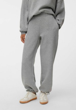 Personne portant un pantalon de jogging gris ample avec des poignets élastiques et des baskets blanches avec des semelles en gomme, debout devant un fond uni.