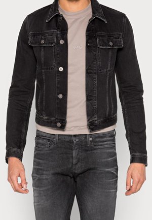 Man draagt een zwarte spijkerjas over een beige T-shirt met de tekst "YPS", gecombineerd met vervaagde zwarte spijkerbroek, armen ontspannen langs het lichaam.