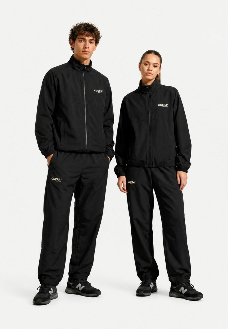 Jeune homme et femme debout côte à côte portant des survêtements noirs assortis avec le logo "CUERA" et des baskets noires, sur un fond blanc uni.