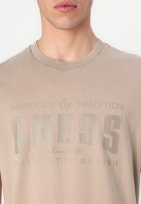 Béžové bavlněné tričko s kulatým výstřihem, krátkými rukávy a výrazným grafickým potiskem s textem "GUESS" a "AUTENTICKÝ DESIGN".
