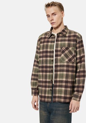 Nuori mies, jolla on päällään ruskea, vihreä ja beige ruudullinen nappipaita valkoisen topin ja sinisten farkkujen päällä, seisoo vaaleaa taustaa vasten.