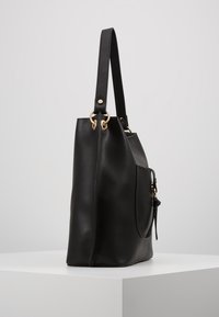 Czarna skórzana torba typu tote z paskiem na ramię i złotymi pierścieniami, stojąca pionowo na białym postumencie na tle jednolitego szarego tła.