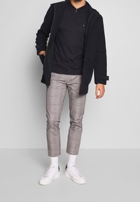 Svart zip-up jacka över en svart polo-skjorta, matchad med rutiga cropped byxor i grått och vita sneakers. Har en ficka fram och ribbade ärmslut.
