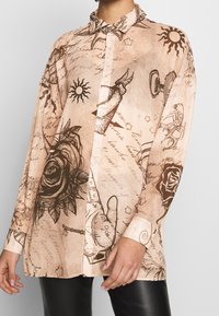 Camicia a maniche lunghe con bottoni in tessuto beige chiaro, caratterizzata da stampe marrone scuro di rose, soli e vari motivi di mani.