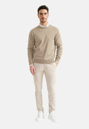Uomo in piedi che indossa un maglione beige sopra una camicia bianca, pantaloni beige e scarpe da ginnastica bianche contro uno sfondo chiaro uniforme.