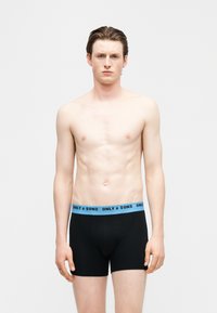 Νέος άνδρας με κοντά καστανά μαλλιά, φορώντας μαύρα μπόξερ με μπλε μέση που φέρει την ετικέτα "ONLY & SONS", στέκεται μπροστά σε απλό φόντο.