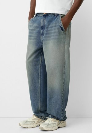 Personne portant un jean bleu bicolore ample, les mains dans les poches et des baskets blanches beige, debout devant un fond blanc uni.