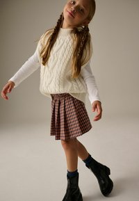 Maglione senza maniche bianco a lavorazione a trecce sopra una camicia a maniche lunghe, abbinato a una gonna a pieghe a quadri marrone e beige e stivali neri alla caviglia.