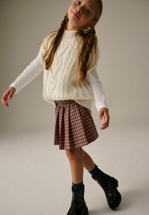 Maglione senza maniche bianco a lavorazione a trecce sopra una camicia a maniche lunghe, abbinato a una gonna a pieghe a quadri marrone e beige e stivali neri alla caviglia.