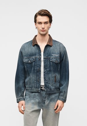 Giovane uomo che indossa una giacca di jeans blu sbiadita con colletto marrone, maglietta bianca e jeans chiari sbiaditi contro uno sfondo semplice.