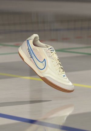 Witte en blauwe Nike Tiempo indoor voetbal schoen zwevend boven een gymzaalvloer met meerkleurige lijnen van het speelveld.