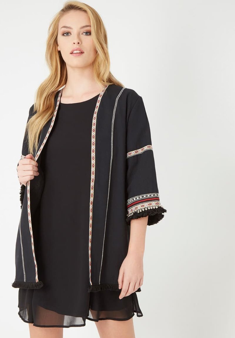 Chaqueta kimono negra con ribete ornamentado en rojo, blanco y dorado, mangas cortas y detalles en flecos texturizados. Acabado de tela suave.