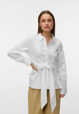 Frau trägt eine weiße Bluse mit Knöpfen, die mit einer Schleife an der Taille gebunden ist, kombiniert mit beigen Hosen, und steht vor einem schlichten hellen Hintergrund.