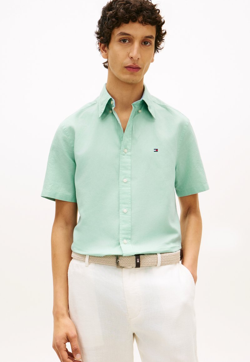 Άνδρας με πουκάμισο κοντομάνικο σε μέντα πράσινο με μικρό λογότυπο, μπεζ υφασμένη ζώνη και λευκό παντελόνι, πάνω σε απλό φόντο.