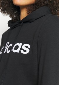 Gros plan d'une personne portant un sweat à capuche noir avec un logo Adidas blanc, les cheveux bouclés visibles au niveau du cou et de l'épaule.