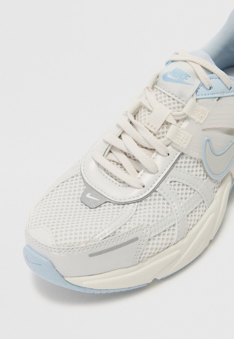 Zapatilla deportiva blanca con parte superior de malla y sintético, acentos en azul claro, lengüeta acolchada y suela texturizada. Presenta un logotipo prominente en el lado.
