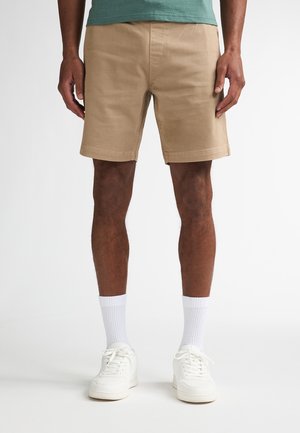 Kort broekje van cognackleurige katoen met rechte pasvorm, voorzien van zijzakken, een gladde textuur en een zoom die net boven de knie valt. Draagt wit sneakers.