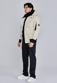 Giacca a vento beige, con colletto alto, molteplici tasche e zip con patch logo. Abbinata a pantaloni neri e sneakers bianche.