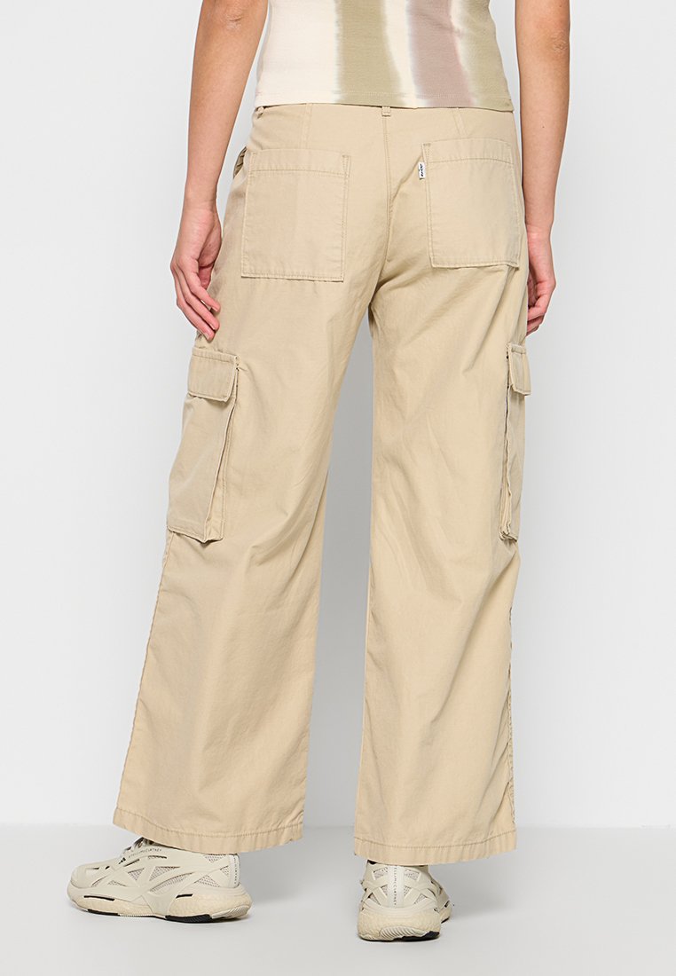 Pantalones cargo beige con piernas anchas, múltiples bolsillos laterales y un bolsillo trasero con cremallera. Confeccionados con un tejido ligero y de textura suave.