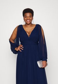 Marineblaues langen Kleid mit tiefem V-Ausschnitt und gefälteltem Oberteil. Hat durchsichtige lange Ärmel und einen glatten Taillenbund. Hält eine silberne Clutch.
