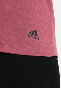 Punainen heather-kankainen t-paita pehmeästä materiaalista, jossa on musta Adidas-logo vasemmassa alareunassa. Lyhyet hihat ja normaali malli.