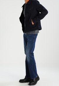 Chaqueta navy con un bolsillo con cremallera, camisa gris debajo, jeans azules y botas negras. La chaqueta tiene un cuello y un corte relajado.