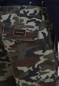 Kamelotervett shorts i grønn, brun og svart stoff; har en baklomme med en brun etikett som det står "Quiksilver" på.