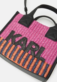 Malá tkaná kabelka s růžovými, oranžovými a modrými pruhy, černým koženým lemováním a velkým černým nápisem "KARL" na přední straně.