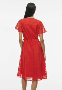 Rochie roșie, texturată, cu mâneci scurte, evazate, un cordon la talie și o fustă până la genunchi, având un model discret în formă de zigzag.