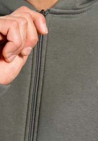 Grå zip-up hoodie med tekstureret stof, synlig lynlås, og en hånd der griber fat i lynlåsgrebet. Fokus på stof og design detaljer.