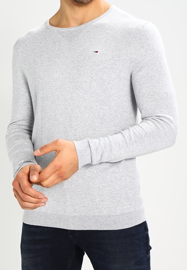 Homme portant un pull léger gris clair à manches longues et col rond, avec un petit logo brodé sur la poitrine, associé à un jean foncé.