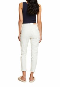 Žena s dlouhými tmavými vlasy, oblečená v námořnickém bezramínkovém topu, bílých džínách s vysokým pasem a béžových nazouvacích sandálech, stojící s výhledem od sebe.