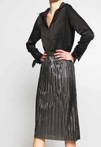 Blouse en satin noir avec col et deux poches avant, associée à une jupe plissée métallique, avec des rayures verticales et une ceinture.