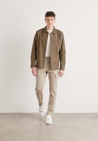 Olivegrön skjortjacka med en framficka, tillsammans med ljusbeige cargobyxor och vita sneakers, stående mot en neutral bakgrund.