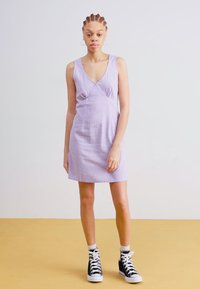 Lilac spaghetti strap dress with a fitted silhouette, textured fabric, and a subtle twist detail at the bust, paired with black high-top sneakers.