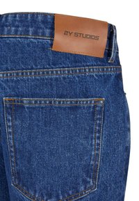 Denim-farkut tummansinisessä värissä, joissa on ruskea nahkainen label vyötäröllä ja "2Y STUDIOS" -teksti sekä näkyvät ompeluaksentit.