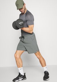 Homem atlético segurando um haltere preto, vestindo uma camiseta cinza ajustada e calções olivados, complementados com sapatos Nike pretos e meias brancas.