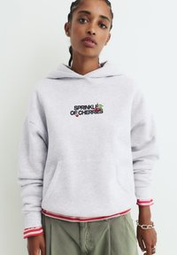 Felpa grigia con cappuccio, con il testo "SPRINKLE OF CHERRIES" e grafica sul petto. Presenta una tasca frontale e polsini a strisce rosse.