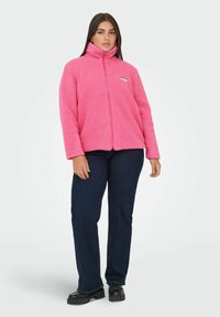 Różowa kurtka z polaru z wysokim kołnierzem, zapięciem na suwak i przednimi kieszeniami. Do tego ciemnoniebieskie dżinsy i czarne buty na platformie.