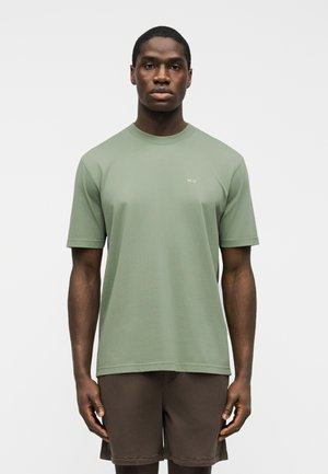 Άνδρας που φοράει κοντομάνικη μπλούζα ανοιχτού πράσινου χρώματος με μικρό λευκό κείμενο και σκούρα καφέ σορτς, στέκεται μπροστά σε απλό λευκό φόντο.