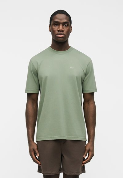 Hombre con camiseta de manga corta verde claro con texto blanco pequeño y pantalones cortos marrón oscuro, de pie contra un fondo blanco liso.