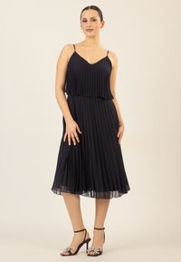 Plissiertes Midikleid in Marineblau, mit verstellbaren Spaghettiträgern und einem geschichteten Oberteil. Es hat einen fließenden Rock und wird mit schwarzen, hochhackigen Sandalen kombiniert.