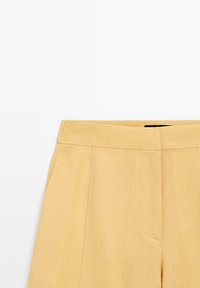 Pantalon beige taille haute avec une couture frontale nette et une fermeture éclair dissimulée sur fond blanc.