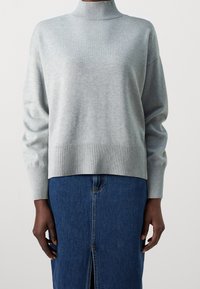 Lichtgrijze trui met een geribde hoge hals, oversized pasvorm en geribbelde zoom. Draagt een donkeren spijkerrok met een voorsplit.