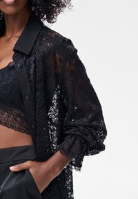Czarna koszula z koronki z kołnierzykiem, ozdobiona guzikami i długimi rękawami. Teksturowana tkanina z kwiatowym wzorem i przezroczystym wyglądem.