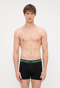 Boxer shorts noirs avec une ceinture élastique verte portant le texte "Originals Studio" en blanc. Texture de tissu lisse, design ajusté.