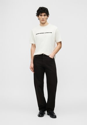 Ung mand med krøllet hår iført hvid Emporio Armani T-shirt, sorte bukser og sorte sko, stående med den ene hånd i lommen.
